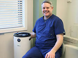 Dr. King smiling while seated next to a medical-grade air purifier in a treatment room
